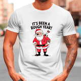 White t-shirt with a graphic of Santa Claus holding an axe and text 'It's Been a Rough Year!' on a blurred street background.