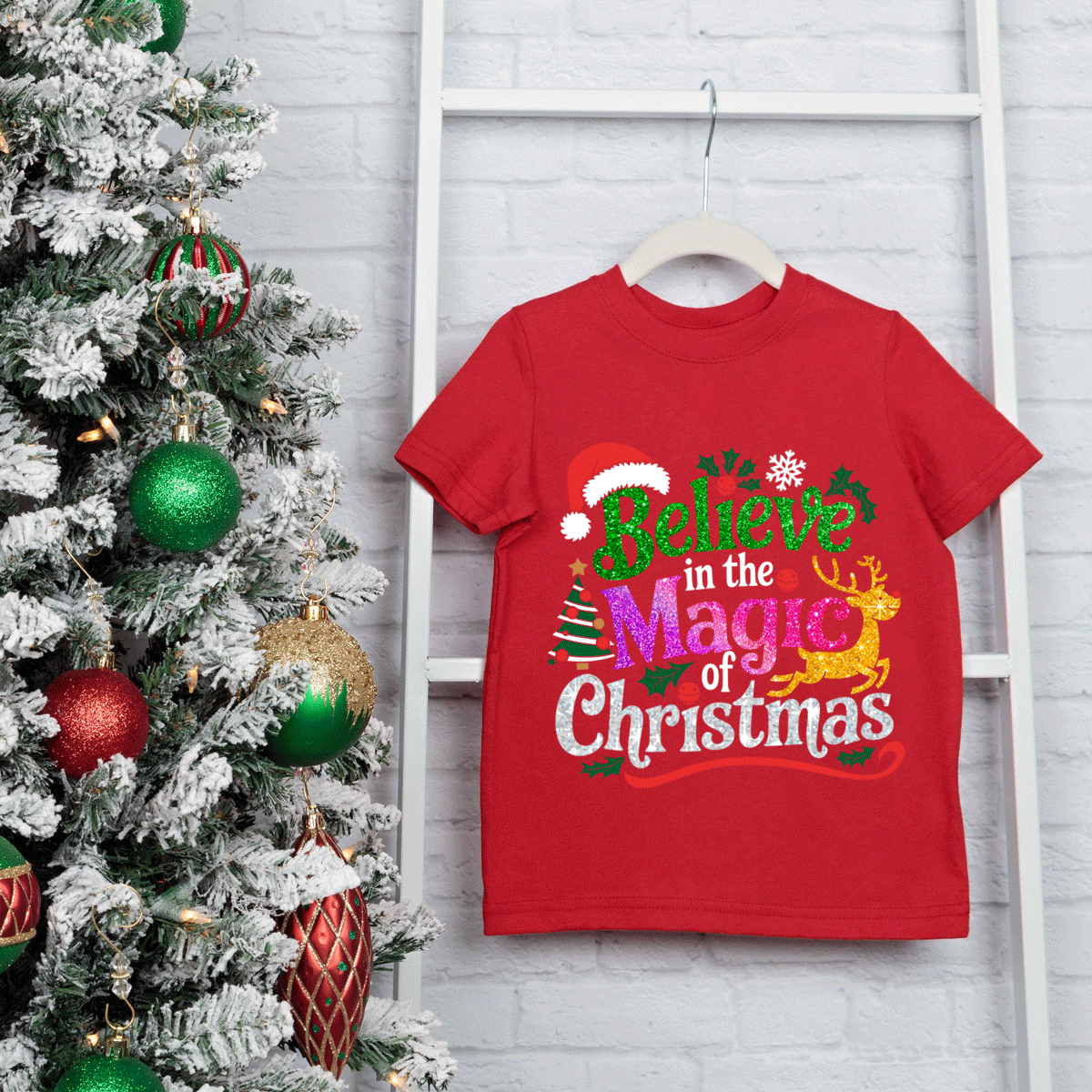 Red t-shirt with Christmas-themed text next to a decorated Christmas tree on a light gray wooden background
