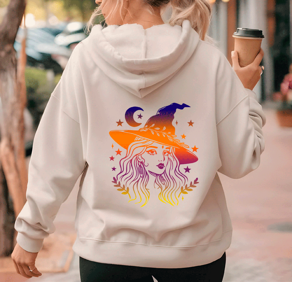 Person wearing a hoodie with a colorful witch graphic on the back, holding a coffee cup.