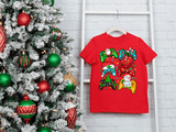 Red t-shirt with Christmas-themed design next to a decorated Christmas tree on a white brick wall.