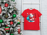 Red t-shirt with Santa Claus design next to a decorated Christmas tree on a white brick wall.