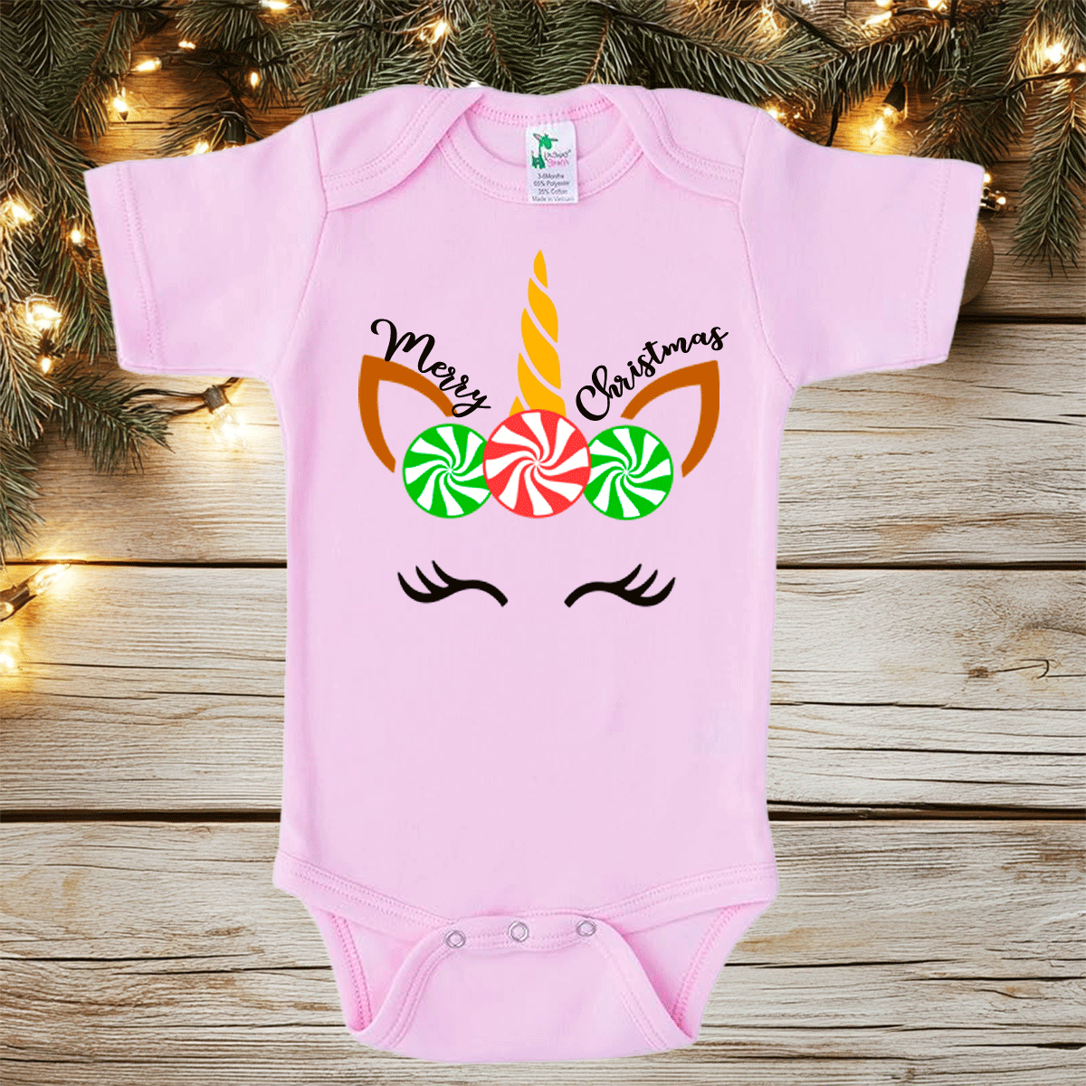Pink baby onesie with unicorn design and candy cane text on a wooden surface with Christmas lights.
