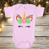 Pink baby onesie with unicorn design and candy cane text on a wooden surface with Christmas lights.