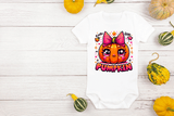 White baby onesie with a pumpkin design and text, surrounded by small pumpkins on a wooden surface.