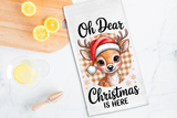 Tea towel with a Christmas-themed design of a deer wearing a Santa hat, text 'Oh Dear Christmas is Here', on a wooden surface with lemons.