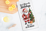 Tea towel with Santa Claus design and humorous text on a kitchen counter with lemons and a cutting board.