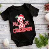 Black baby onesie with 'My 1st Christmas' print featuring a Santa hat on a wooden surface with Christmas decorations.