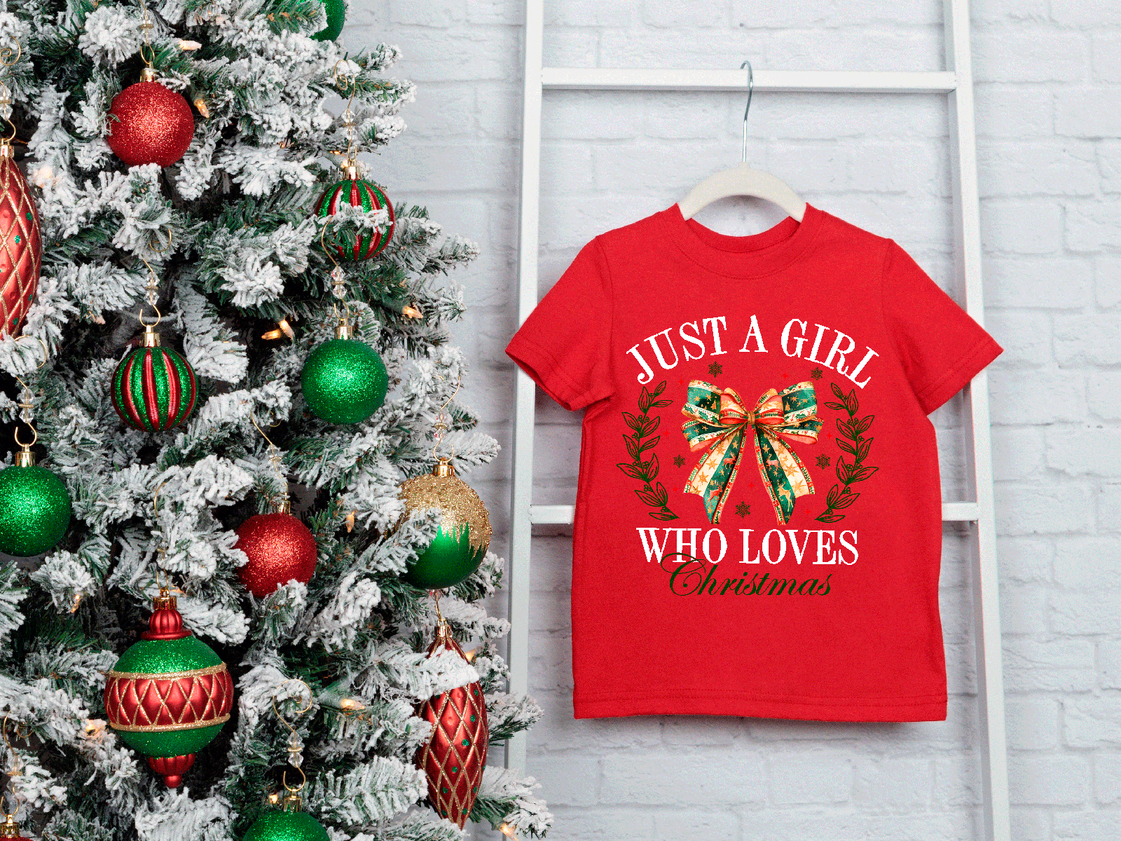 Red t-shirt with Christmas-themed design next to a decorated Christmas tree on a white brick wall.