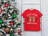 Red t-shirt with Christmas-themed design next to a decorated Christmas tree on a white brick wall.