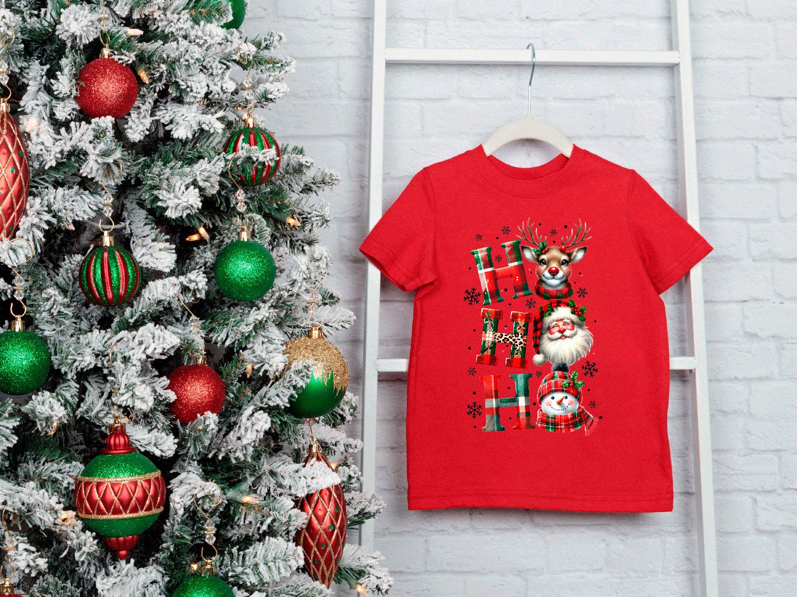 Red t-shirt with Christmas design next to a decorated tree on a white brick wall.