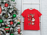 Red t-shirt with Christmas design next to a decorated tree on a white brick wall.