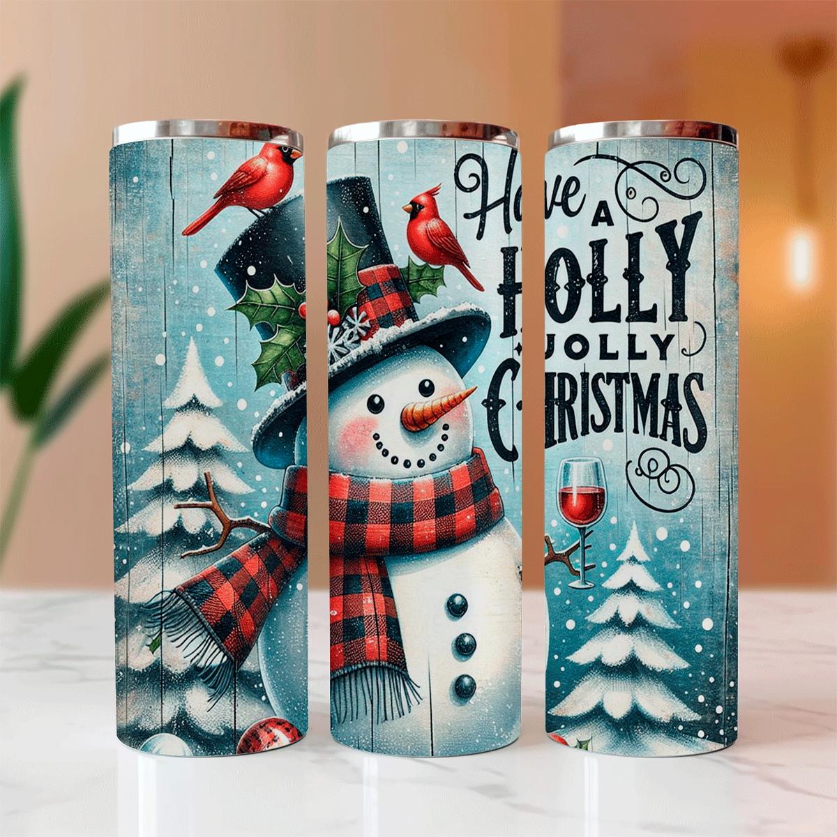 Three Christmas-themed tumblers with snowmen, cardinal birds, and festive text on a blurred indoor background.