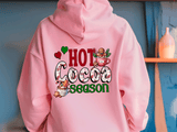 Pink hoodie with 'Hot Cocoa Season' text and graphics on a blue wall background