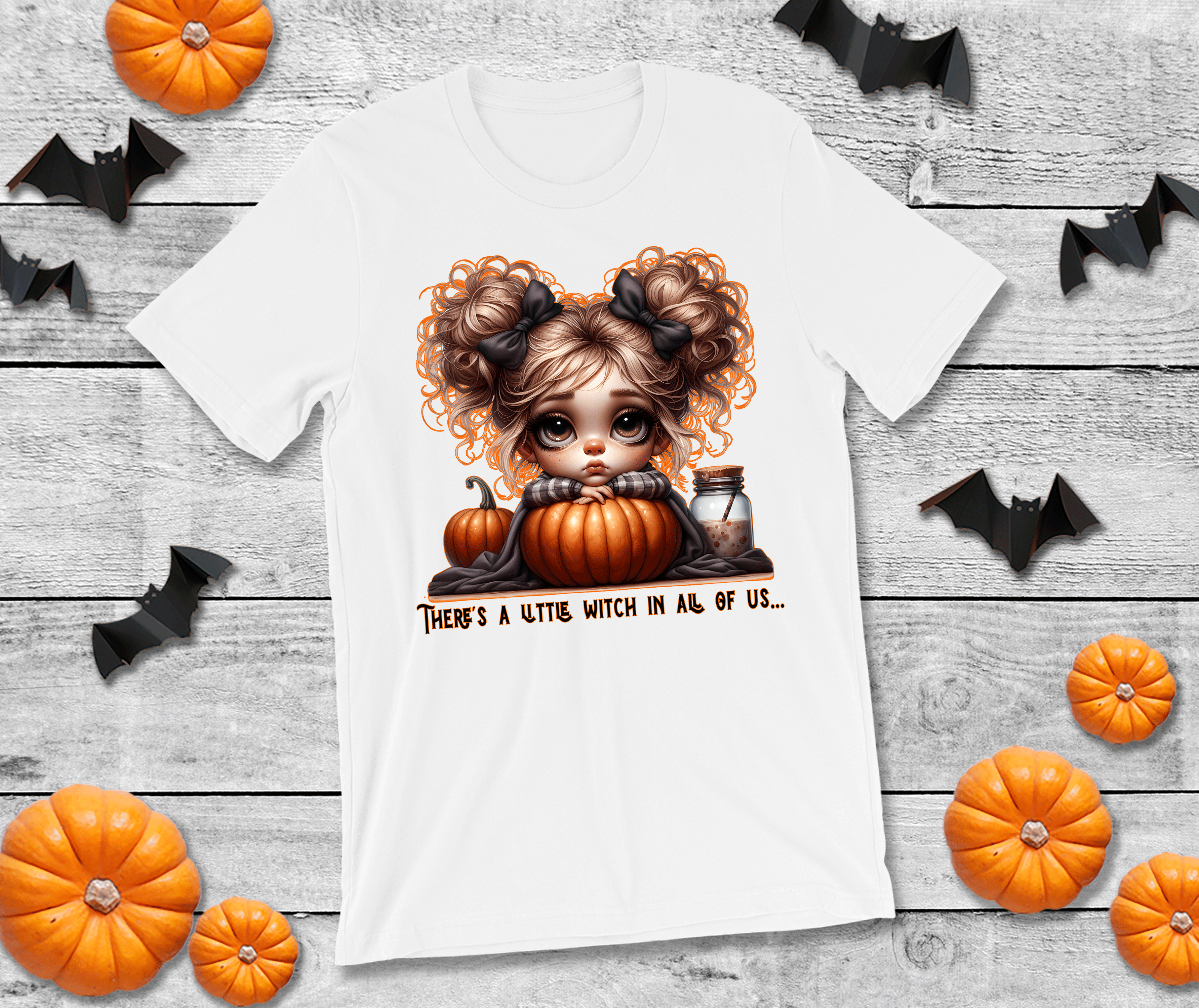 White t-shirt with a Halloween-themed design of a witch and pumpkins on a wooden surface with decorative bats and pumpkins.