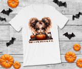 White t-shirt with a Halloween-themed design of a witch and pumpkins on a wooden surface with decorative bats and pumpkins.