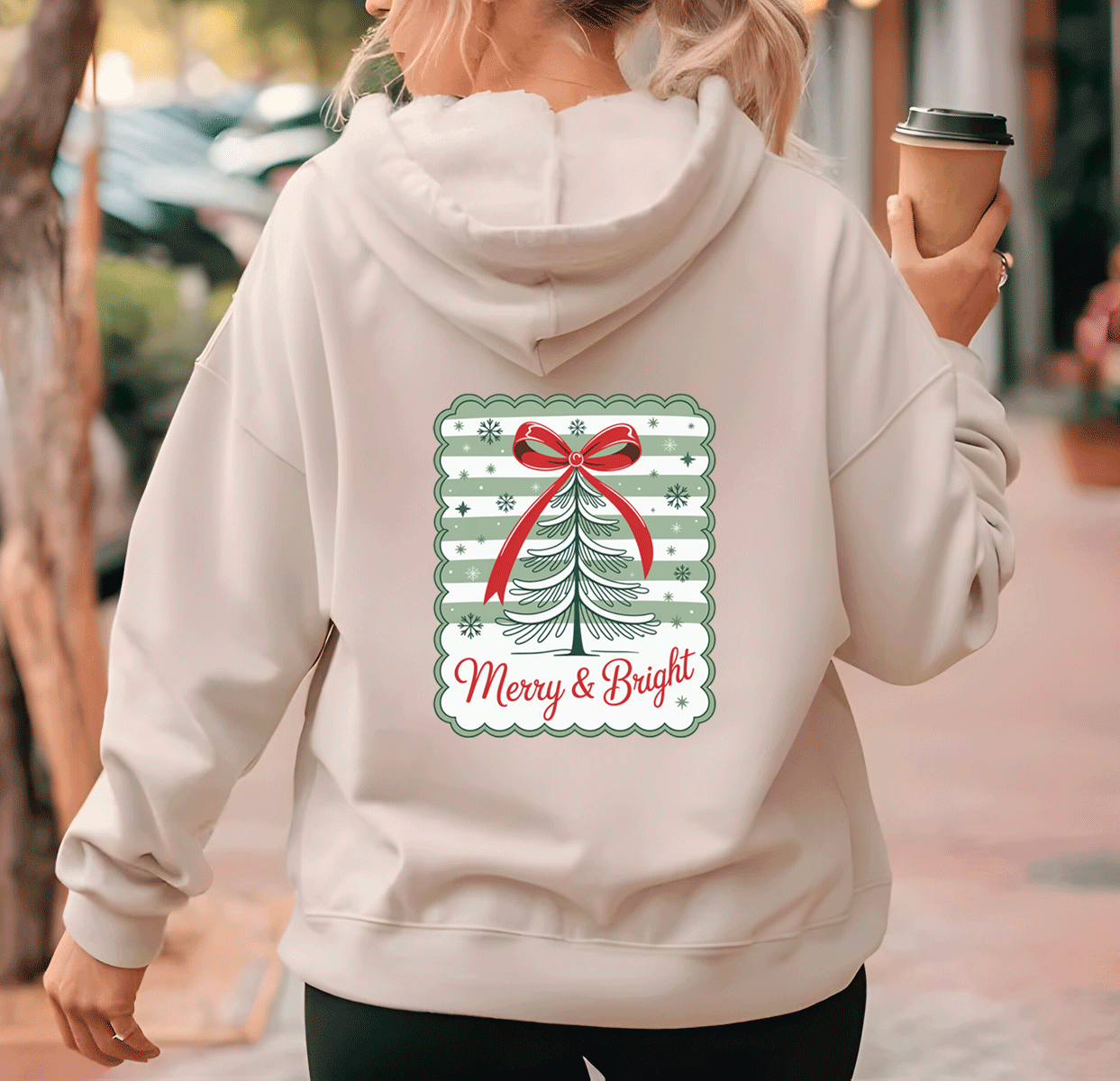 Person wearing a hoodie with a Christmas design and text, holding a coffee cup.