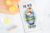 Tea towel with a snowman design and text 'Mix and Mingle' on a kitchen counter with lemons.