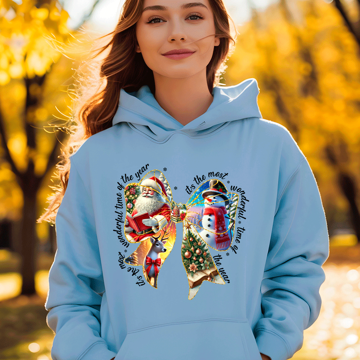 Woman wearing a light blue hoodie with Christmas-themed designs and text, standing in an autumn setting.