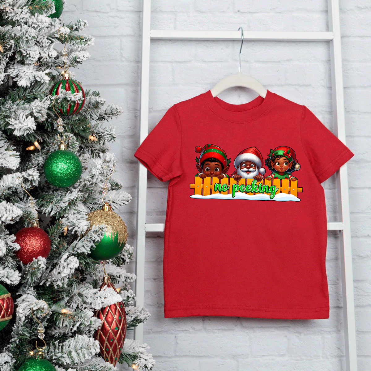 Red t-shirt with Christmas-themed design on a hanger next to a decorated Christmas tree.