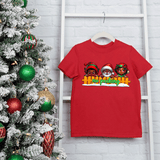 Red t-shirt with Christmas-themed design on a hanger next to a decorated Christmas tree.