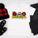 White t-shirt with Christmas-themed design featuring Santa Claus and two children, surrounded by a beanie, slippers, and jeans on a white background.
