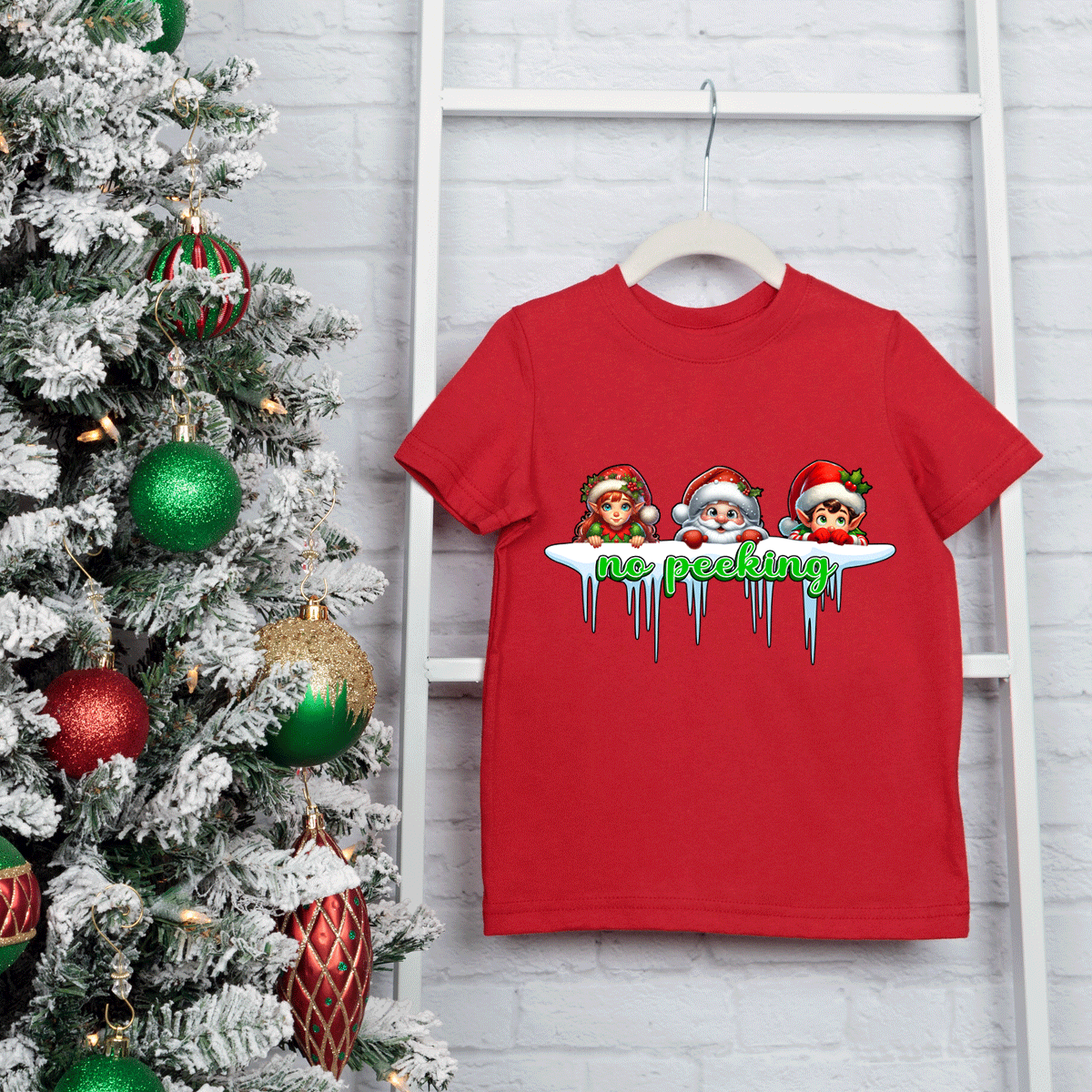 Red t-shirt with Christmas-themed design hanging on a ladder next to a decorated Christmas tree.