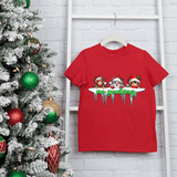 Red t-shirt with Christmas-themed design hanging on a ladder next to a decorated Christmas tree.