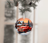 Christmas ornament with a red truck and snowy village scene, hanging from pine branches.