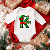 White baby onesie with a green and red design on a red fabric background with Christmas decorations.