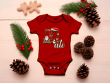 Red baby onesie with Christmas-themed design on a wooden surface with pine cones and greenery.