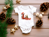 White baby onesie with Christmas design and text on a wooden surface with decorative items.
