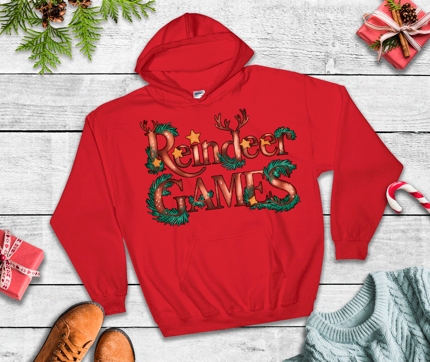 Red hoodie with 'Reindeer Games' text on a wooden surface with Christmas decorations.