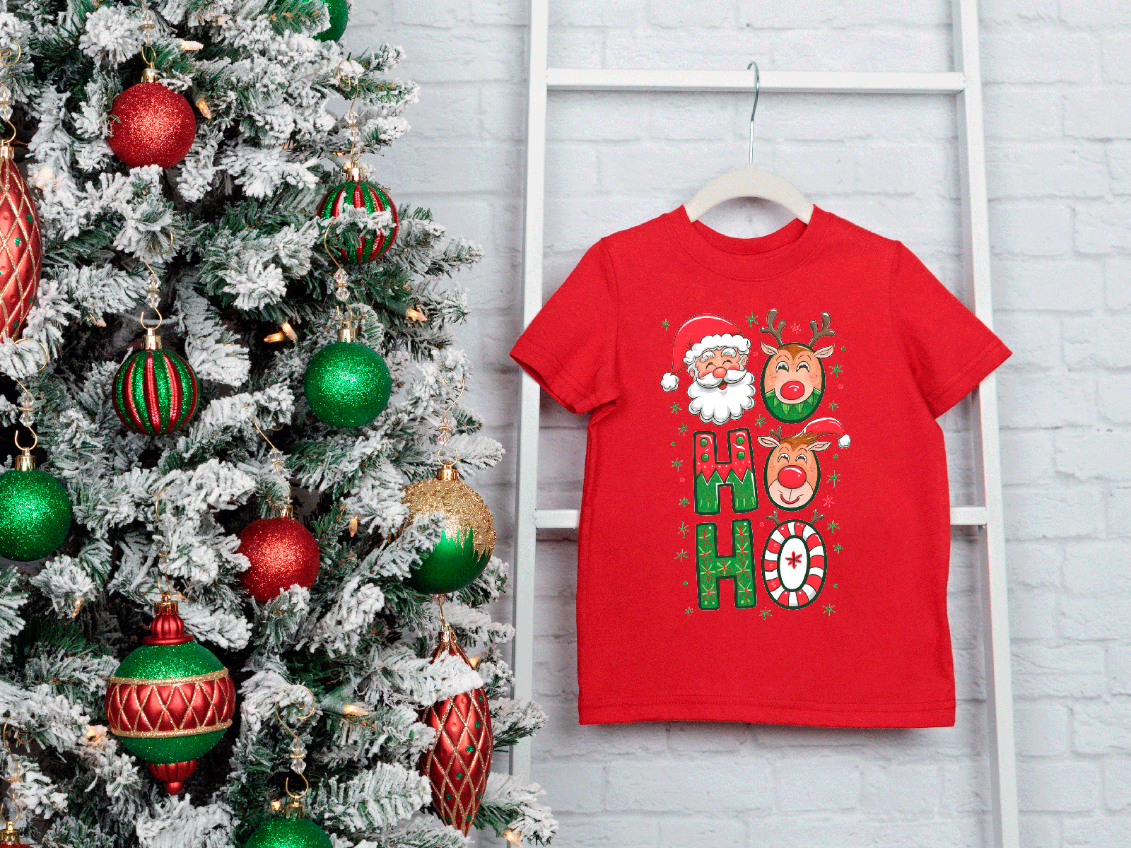 Red t-shirt with Christmas design next to a decorated tree on a white brick wall.