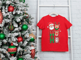 Red t-shirt with Christmas design next to a decorated tree on a white brick wall.