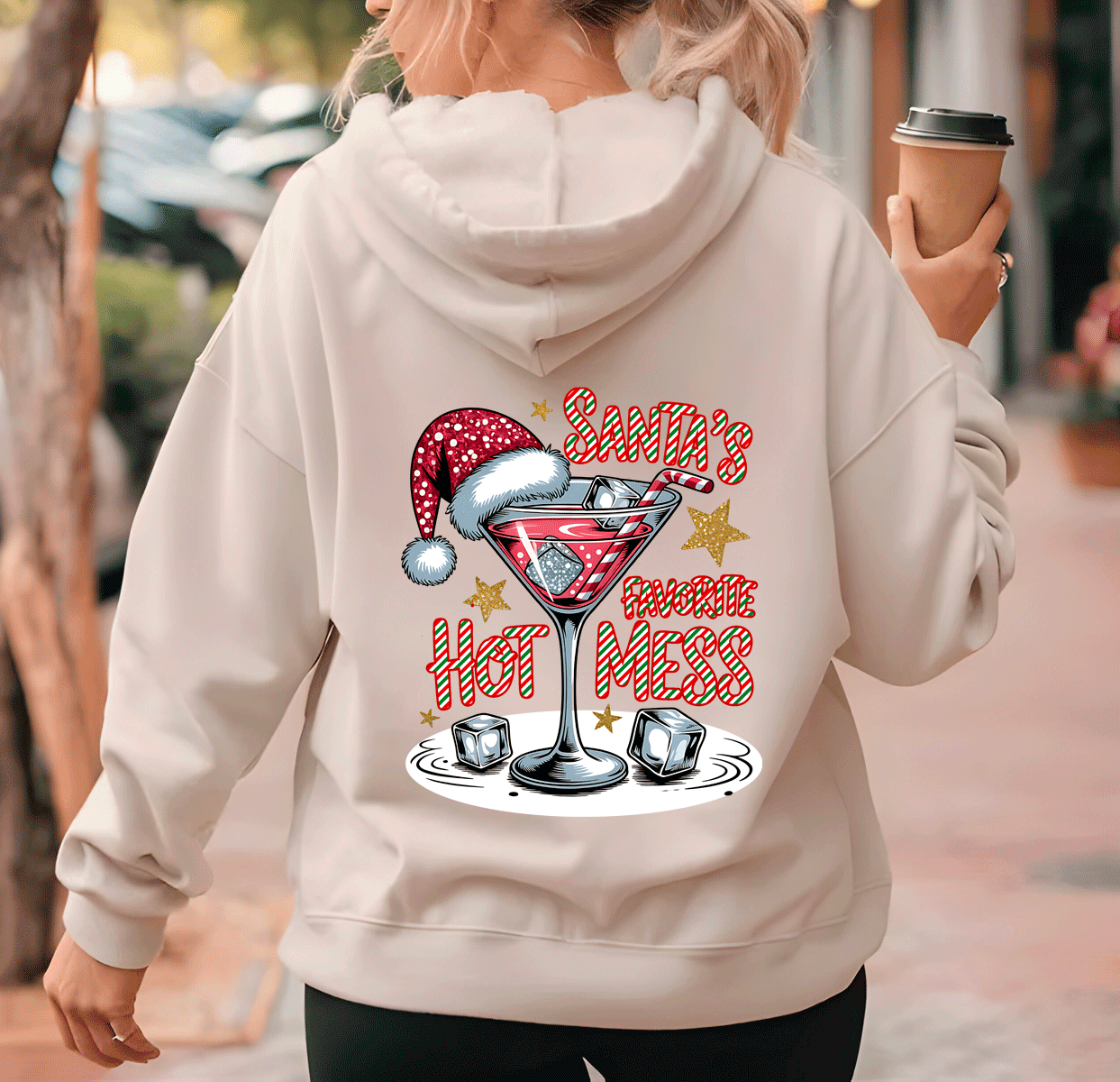 Person wearing a hoodie with a Christmas-themed design of a martini glass and text, holding a coffee cup.