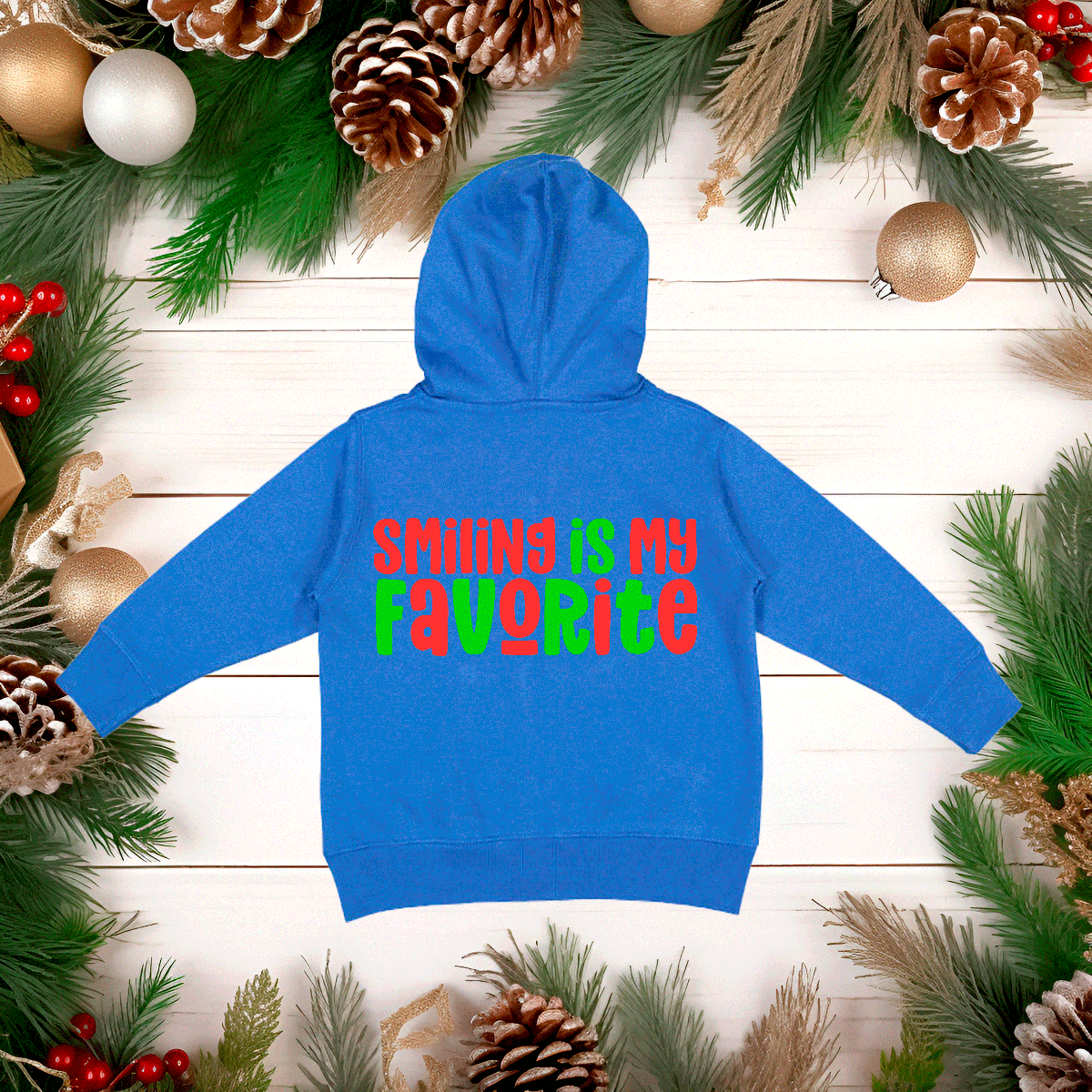 Blue hoodie with 'SMILING IS MY FAVORITE' text on a wooden surface with Christmas decorations.