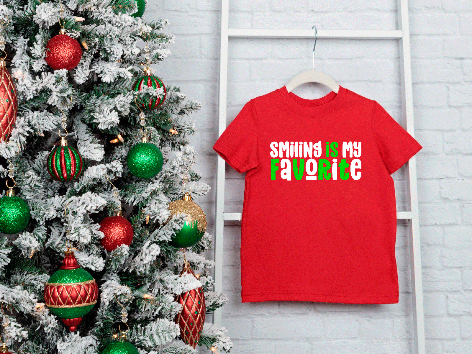 Red t-shirt with 'SMILING IS MY FAVORITE' text next to a decorated Christmas tree on a white brick wall.