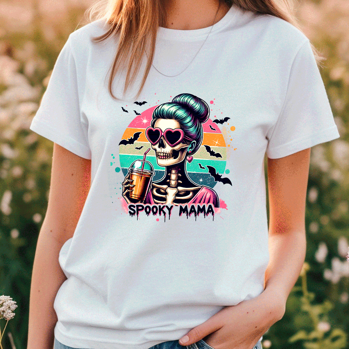 Person wearing a white t-shirt with a 'Spooky Mama' skeleton design in a field.