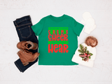 Green t-shirt with Christmas-themed text, jeans, boots, and a decorative bag on a light background