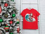 Red t-shirt with Santa Claus surfing design next to a decorated Christmas tree.