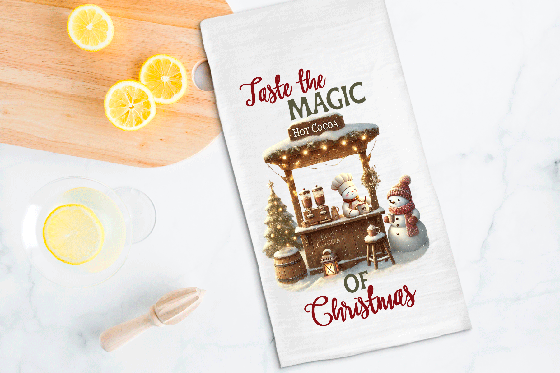 Tea towel with Christmas design featuring a hot chocolate stand and snowman, placed on a table with lemon slices and a wooden spoon.
