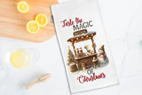 Tea towel with Christmas design featuring a hot chocolate stand and snowman, placed on a table with lemon slices and a wooden spoon.