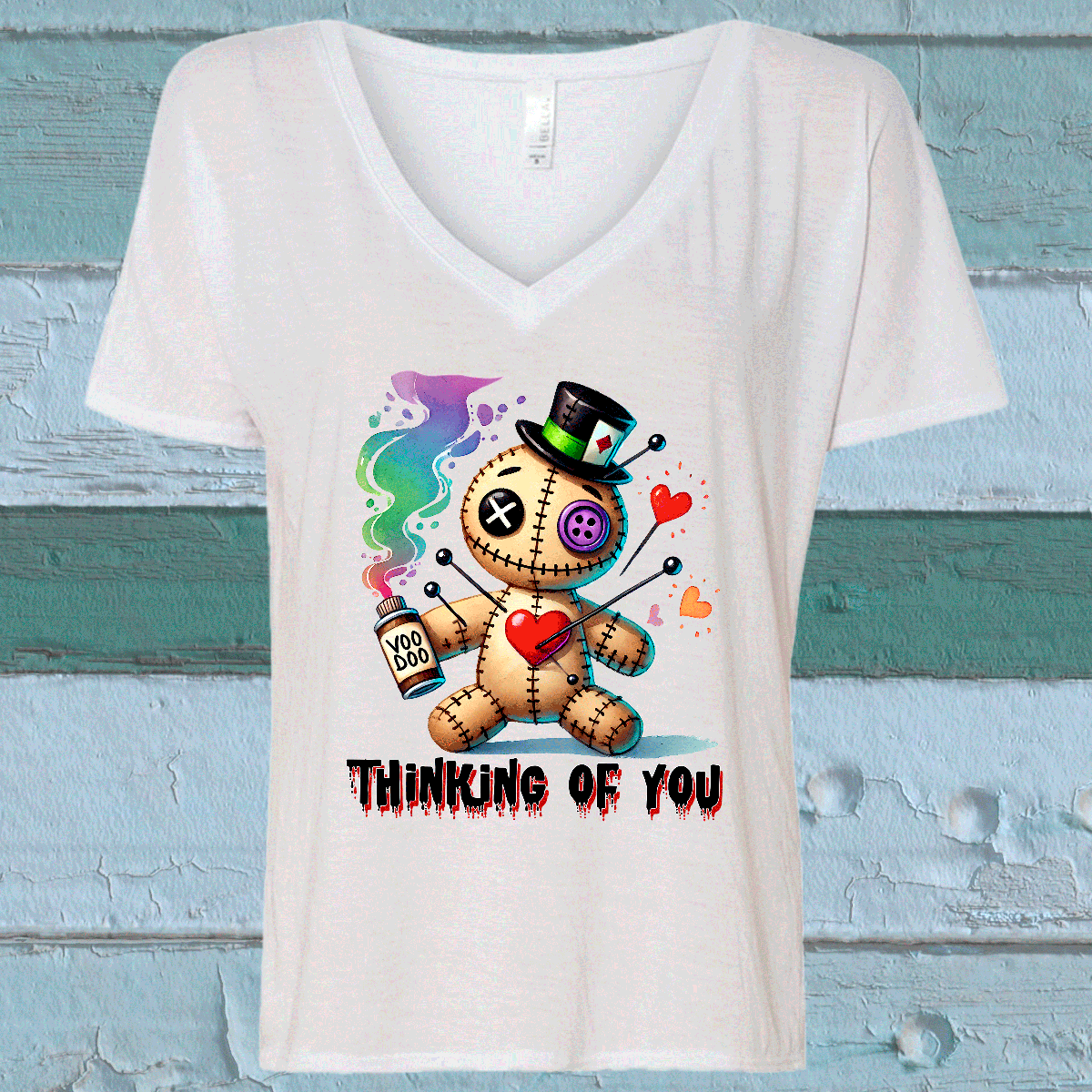 White V-neck t-shirt with a colorful graphic of a teddy bear holding a heart and a bottle, with text 'Thinking of You' on a wooden background.
