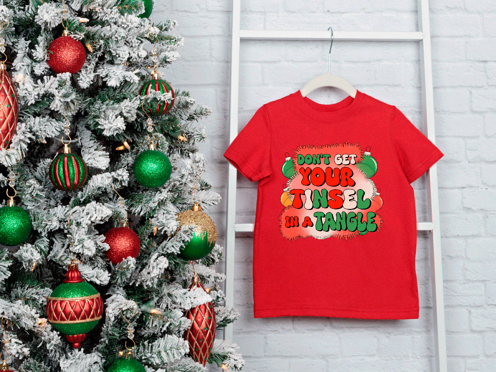 Red t-shirt with Christmas-themed design next to a decorated Christmas tree on a white brick wall.
