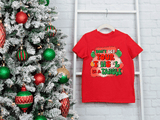Red t-shirt with Christmas-themed design next to a decorated Christmas tree on a white brick wall.