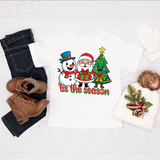 Christmas-themed shirt with Santa, snowman, and tree design on a white background with jeans, boots, and a beanie.