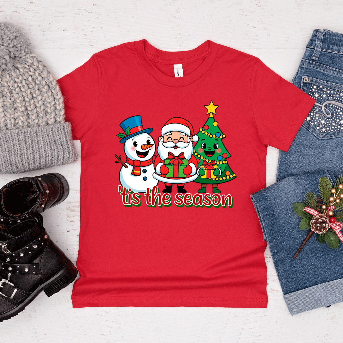 Red t-shirt with Christmas design featuring Santa, snowman, and tree, surrounded by winter accessories on a light background.
