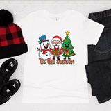 White t-shirt with Christmas design featuring Santa, snowman, and tree, surrounded by festive items on a white background.