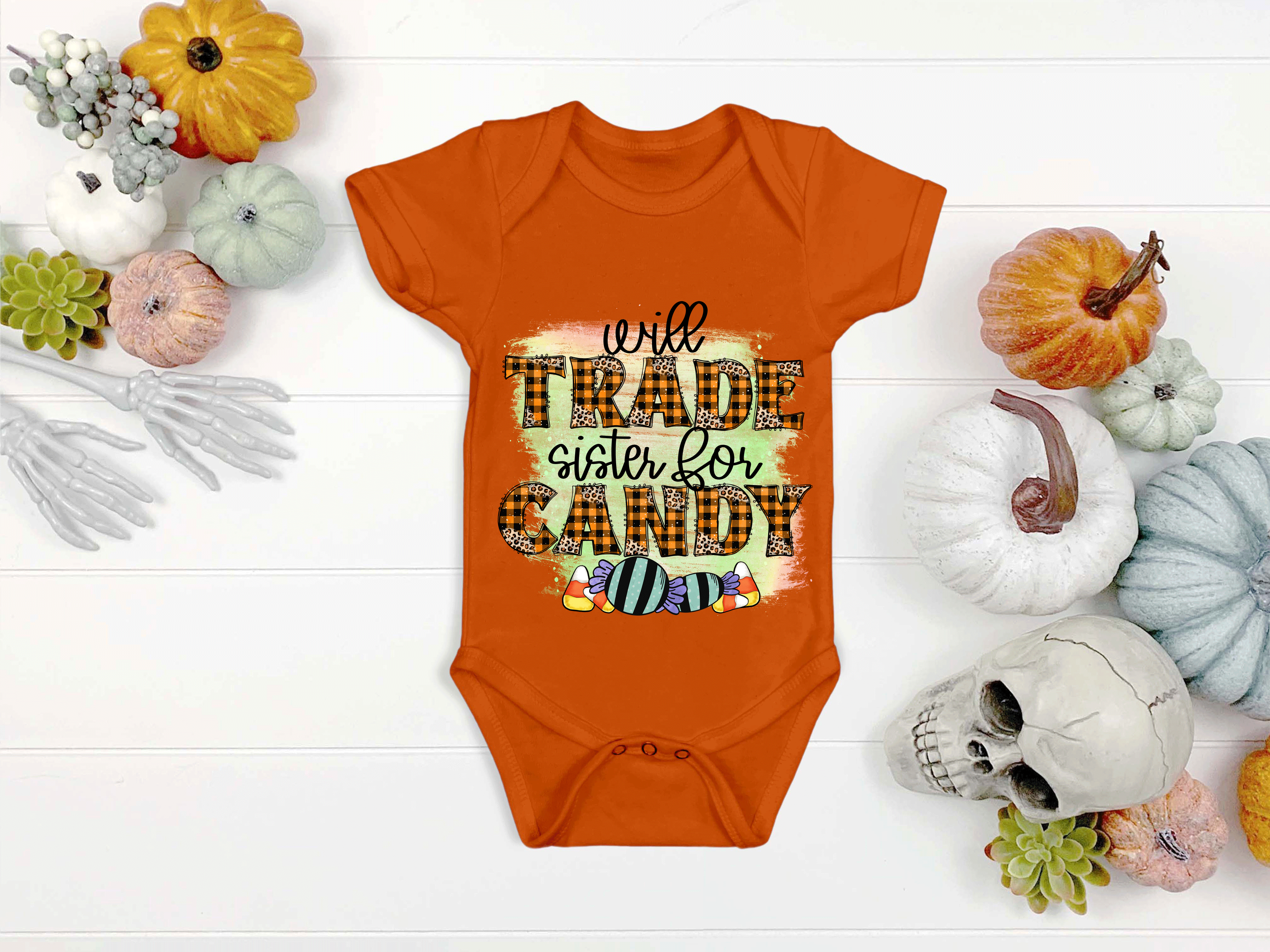 Orange baby onesie with Halloween text on a white surface with pumpkins and decorations.