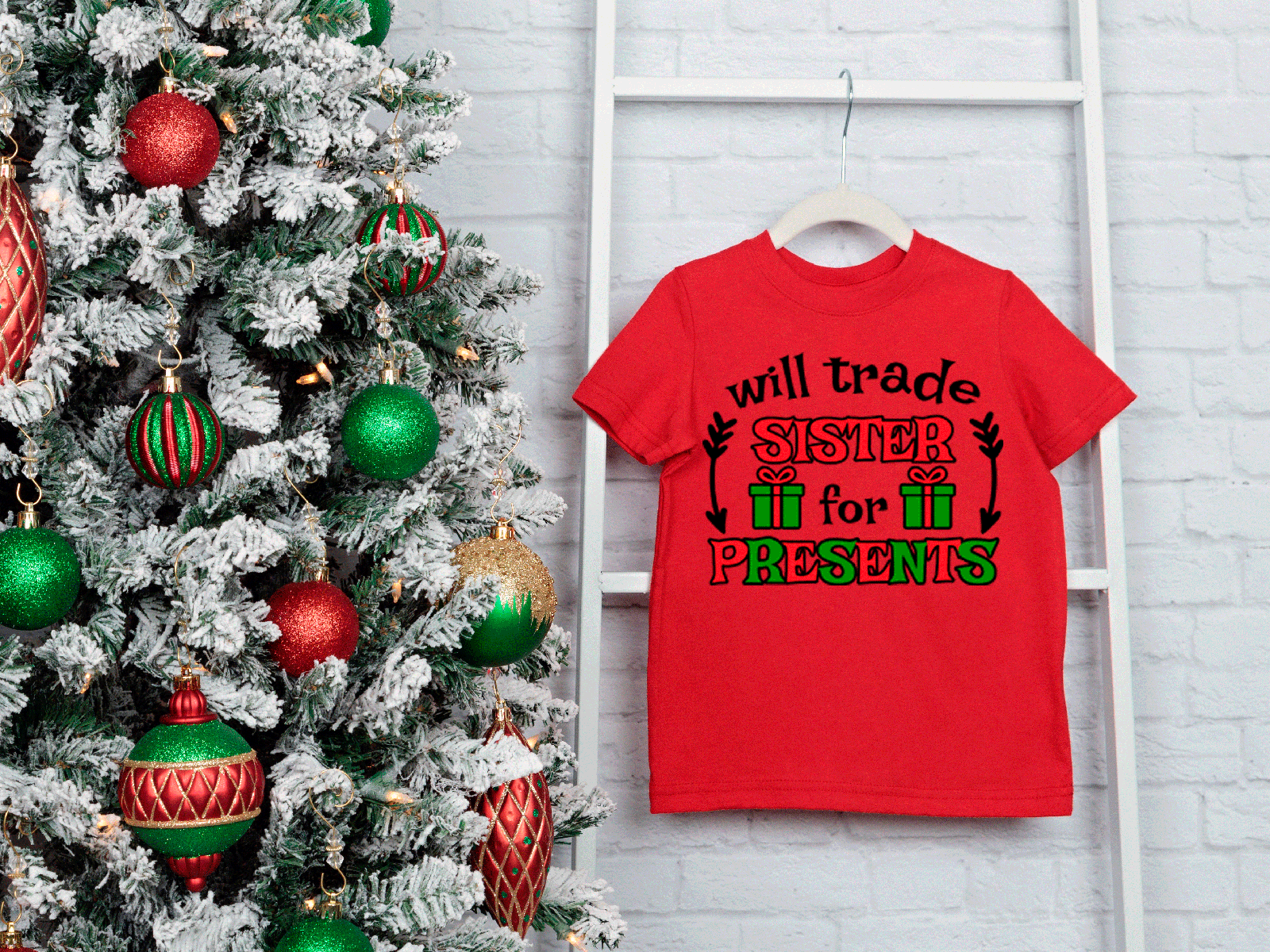Red t-shirt with Christmas-themed text next to a decorated Christmas tree on a white brick wall.